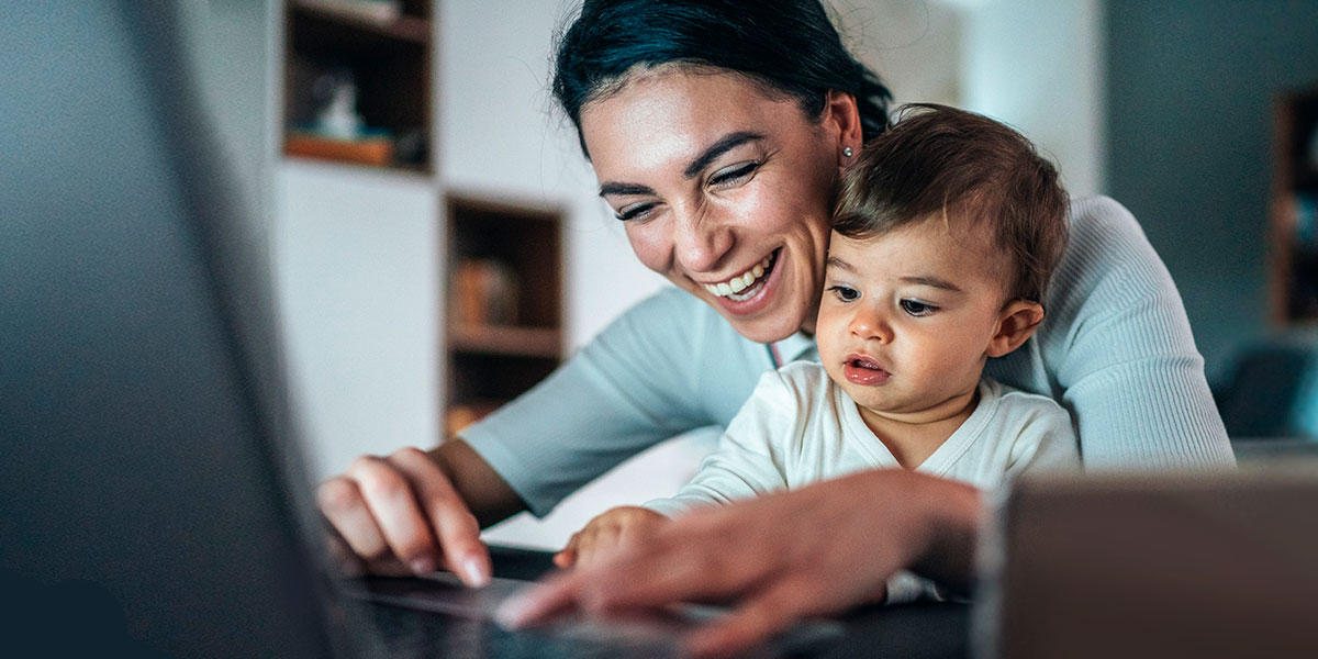 Mother smiling with baby on her lap working on a laptop.
