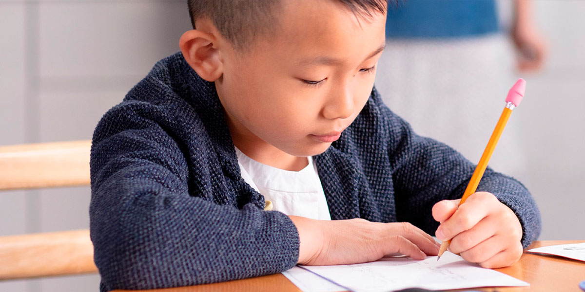 Asian boy writing on paper with a pencil in a classroom.