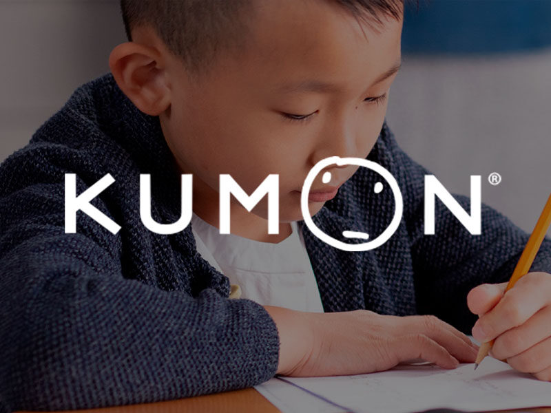Kumon logo over an image of a Asian boy writing on paper with a pencil in a classroom.