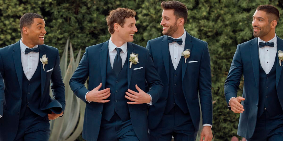 A groom and three groomsmen dressed in navy blue suits walking outdoors.
