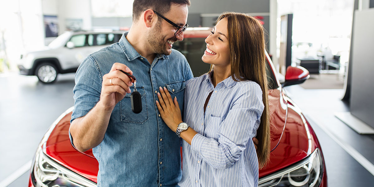 Man holding new car keys with a woman hugging him in front of a red car.