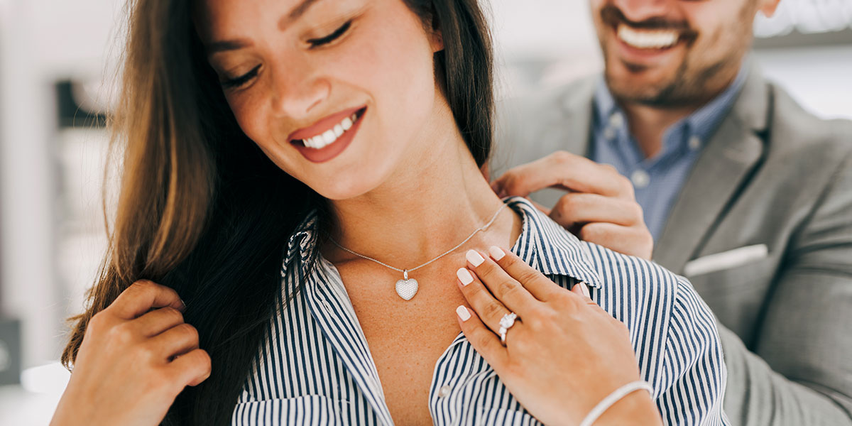 Woman standing with her eyes closed with a man behind her putting a necklace on her.