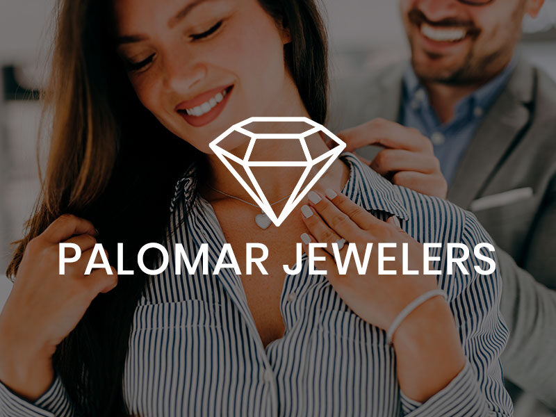Palomar Jewelers logo over an image of a Woman standing with her eyes closed with a man behind her putting a necklace on her.