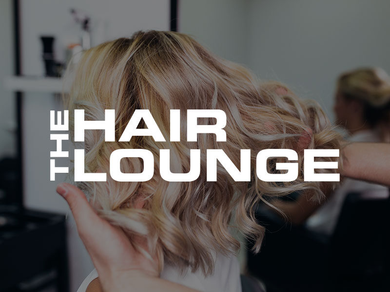 The Hair Lounge logo over an image of a Woman showing off newly colored and curled hair in a salon chair.